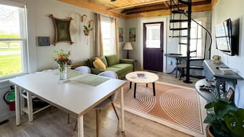 A cozy living room with a wooden ceiling, a dining table, a sofa, a TV, and a staircase leading to another level.