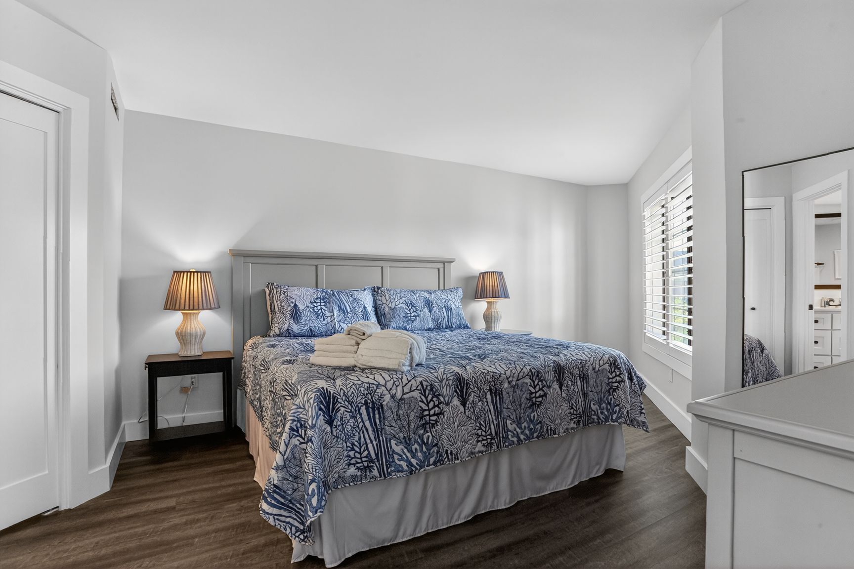 Une chambre à coucher avec un lit, deux tables de chevet avec des lampes, une commode et une fenêtre avec des stores.
