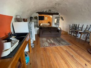 Een interieur met een houten vloer, een bank, een vloerkleed, een eettafel met stoelen en een keuken met een koelkast en een spoelbak.
