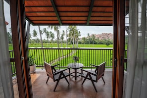 An open balcony with a wooden table and chairs, a potted plant, and a view of a green landscape with palm trees and a rock formation.