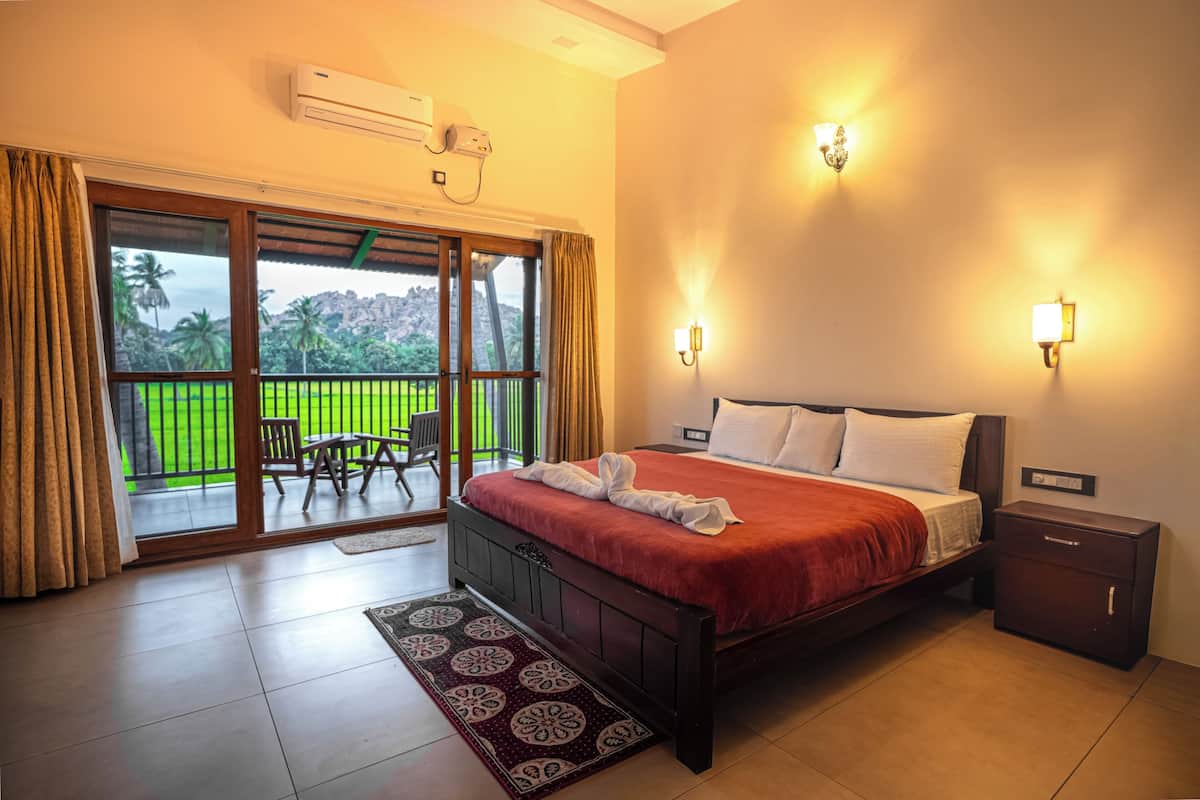 A hotel room with a bed, a nightstand, a wall-mounted air conditioner, a sliding glass door to a balcony, and a view of greenery.