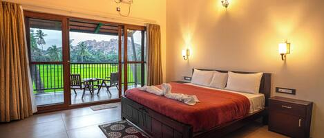 A hotel room with a bed, a nightstand, a wall-mounted air conditioner, a sliding glass door to a balcony, and a view of greenery.