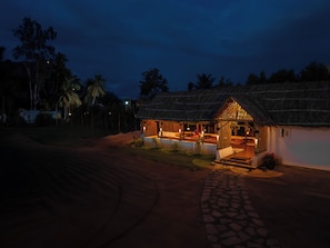 A thatched-roof building with a paved entrance, surrounded by palm trees and a clear sky.