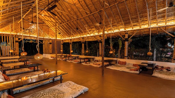 Un espacio interior con techo alto, vigas de madera, lámparas colgantes y alfombras estampadas.