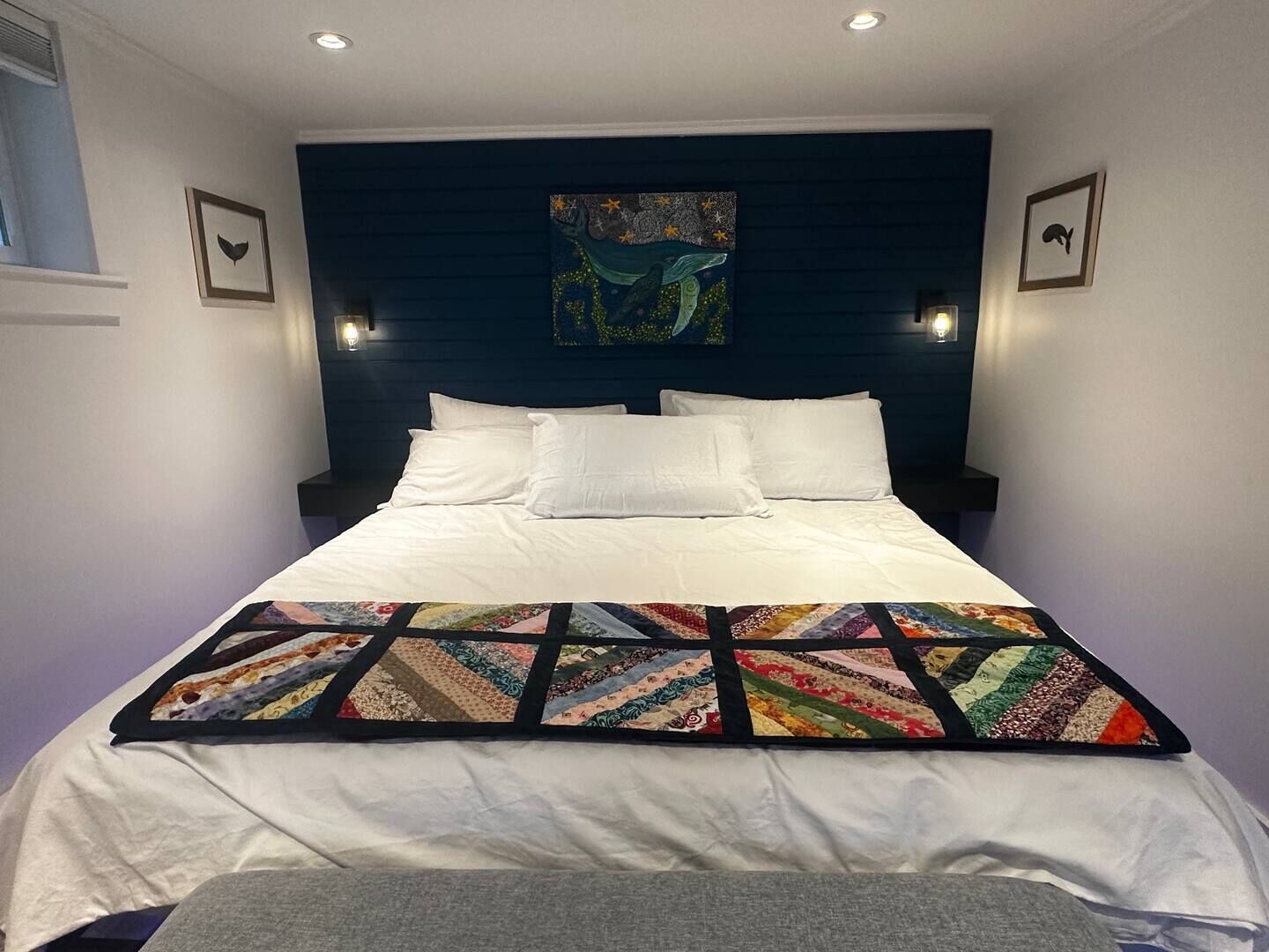 Una cama bien tendida con sábanas blancas y un edredón colorido, un cabecero oscuro y luces fijadas a la pared.