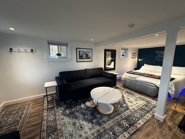 A modern bedroom with a black sofa, a round white coffee table, a bed with a grey headboard, and a large mirror. - The Whistling Kettle | King Studio Near Downtown (St. John's)