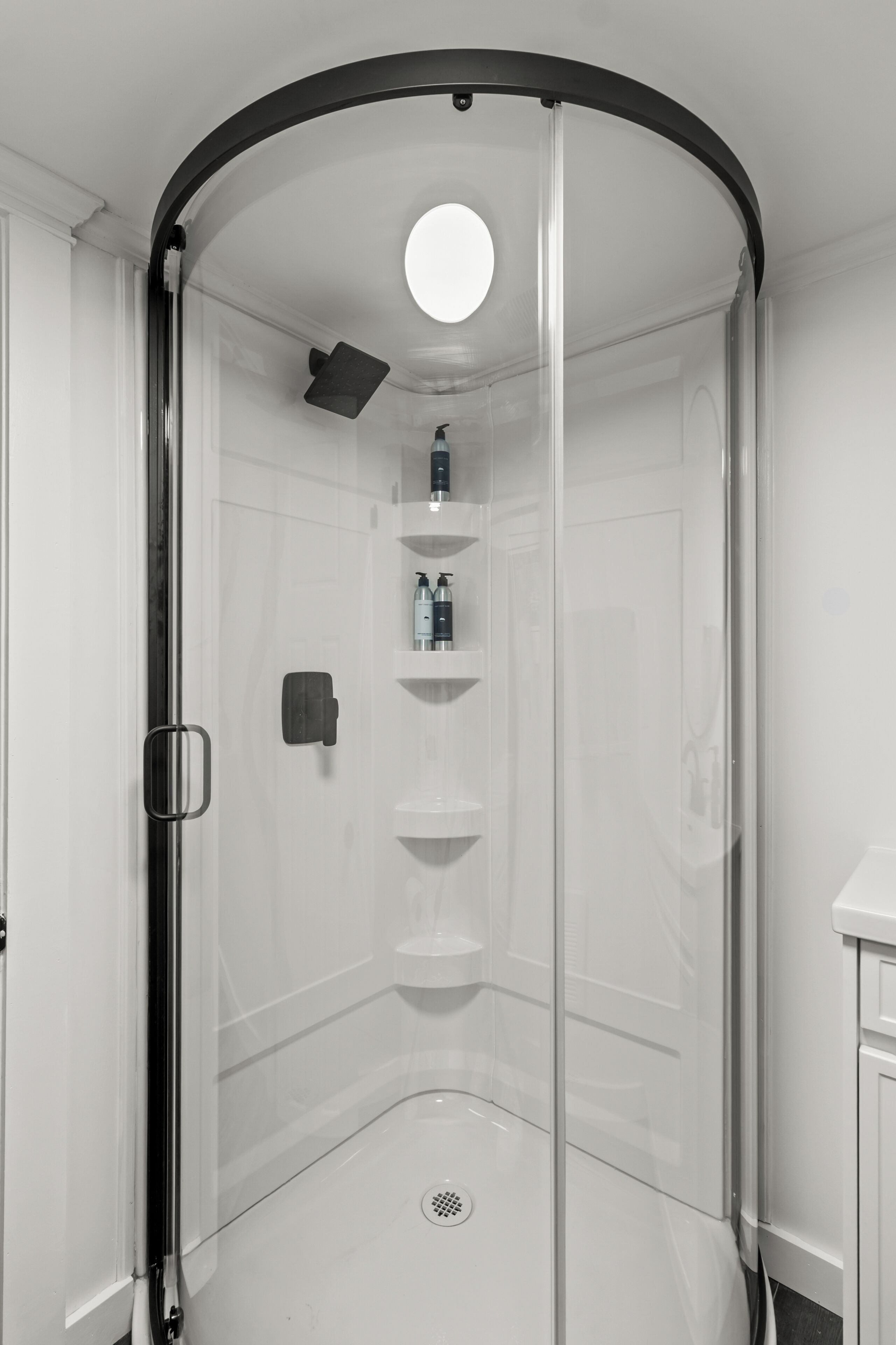 Un baño moderno con ducha de vidrio, una lámpara de techo redonda y piso cubierto de baldosas blancas.