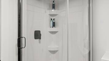 Un baño moderno con ducha de vidrio, una lámpara de techo redonda y piso cubierto de baldosas blancas.