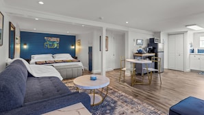 Een moderne woonkamer met een blauwe accentmuur, een bank, een kleine ronde tafel en een keukenhoek met witte kasten en een magnetron.