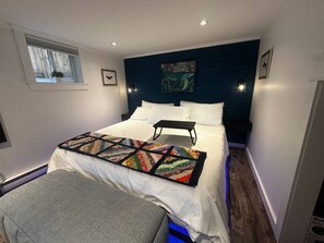 A bedroom with a bed, a bedside table, a window, and a wall-mounted artwork. - The Whistling Kettle | King Studio Near Downtown (St. John's)