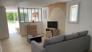 Una sala de estar moderna con un sofá gris, una mesa de centro de madera, un televisor sobre un mueble de madera y un ventanal con vistas a la vegetación.
