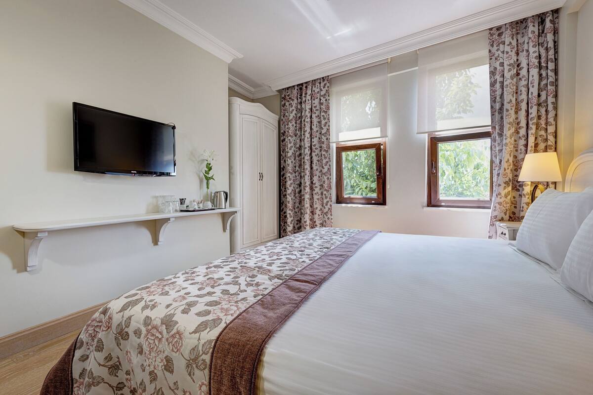 Une chambre d’hôtel avec un lit, un téléviseur fixé au mur, une fenêtre avec des rideaux et une table de chevet avec une lampe.