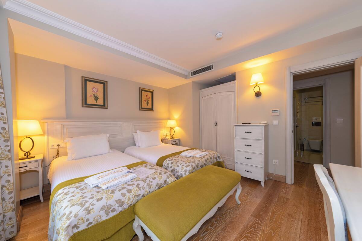 Une chambre d’hôtel avec deux lits, une table de chevet avec une lampe, une commode et une salle de bain visible à travers une porte ouverte.