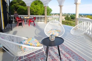A covered outdoor seating area with white wicker chairs and a table, a white railing, and a view of greenery and buildings in the background. - Villa El Dorado – Private Oasis near the Beach (Estepona)