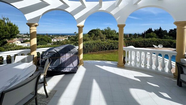 Ein überdachter Außenbereich mit weißen Säulen, einem Grill und Blick auf die Landschaft.