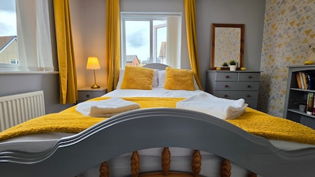 A bedroom with a bed, a bedside lamp, a window with curtains, a dresser, a mirror, and a bookshelf.. Stylish & Modern 4BR Home in Shirebrook