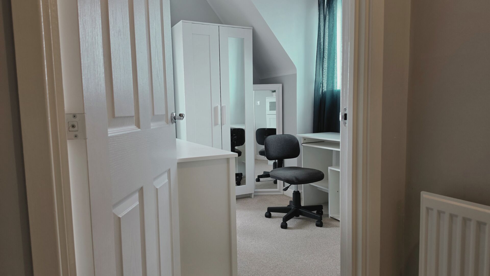 A narrow hallway with a white door, a black office chair, and a white desk with shelves.