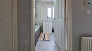 Ein schmaler Flur, der zu einem Badezimmer mit Dusche, Waschbecken und kleinem Fenster führt.