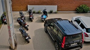 A parking area with several motorcycles and a black SUV, a white car, and a brick wall in the background. - Casa Bella  (Sohna)