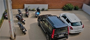 Un área de estacionamiento con varias motocicletas y un SUV negro, un coche blanco, y un muro de ladrillo al fondo.
