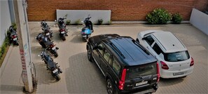 A parking area with several motorcycles and a black SUV, a white car, and a brick wall in the background. - Casa Bella  (gurugram)