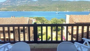 En balkon med hvide stole, udsigt over et kystlandskab med både, og bjerge i baggrunden.