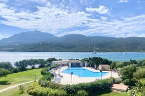 Una piscina rodeada de follaje, un edificio con techo de tejas y una vista de un lago con barcos y montañas al fondo.
