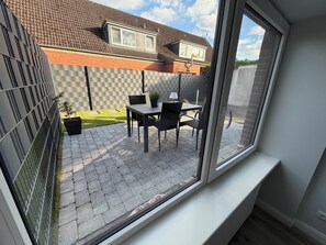 Vista pela janela de um pátio com mesa e cadeiras, um pequeno jardim e uma casa com telhado de telhas.