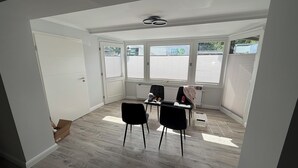 Quarto com porta branca, radiador, duas cadeiras pretas, uma mesa e janela com persianas.