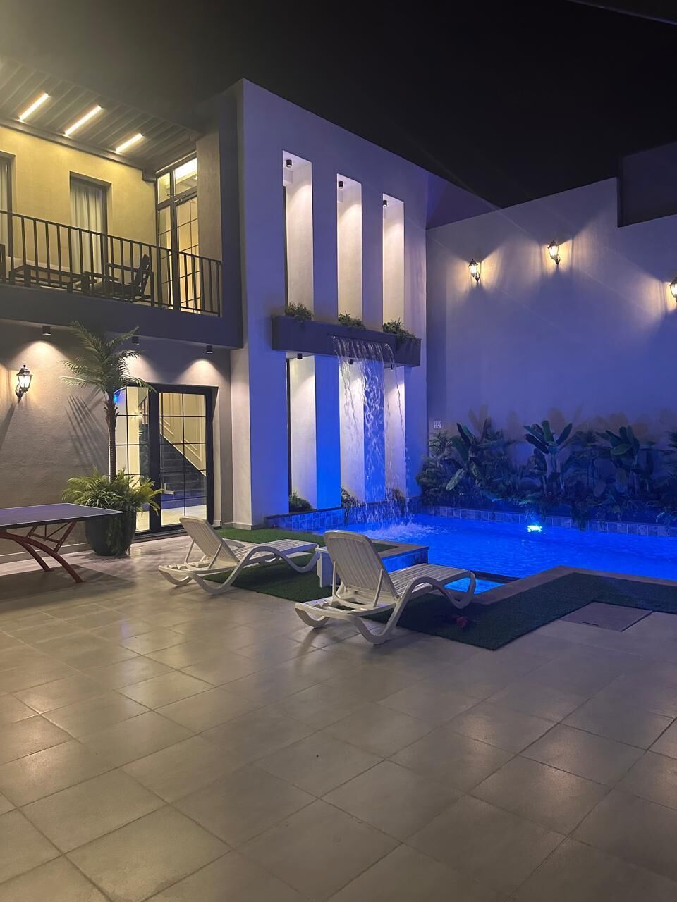 Un espace piscine extérieur moderne, doté de chaises longues, d’une petite cascade et d’un éclairage d’ambiance.