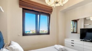 A hotel room with a bed, a television, a window with a view, and a chandelier. - Anastasia Suite of Sun & Sea | Bright & Comfy Cretan Getaway (Vathianos Kampos)