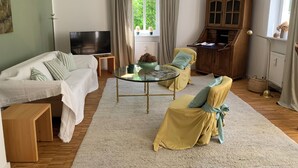 Una sala de estar con un sofá, sillones, una mesa de centro de vidrio, un televisor, un aparador de madera y una ventana con cortinas.