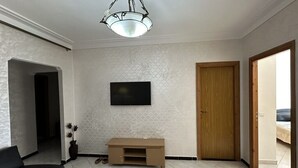Quarto com televisão na parede, porta de madeira e um pequeno móvel para entretenimento.