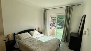 Quarto com cama, mesas de cabeceira, cômoda e uma porta de vidro deslizante para uma varanda.