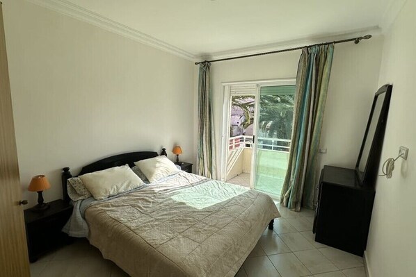 Une chambre à coucher comprenant un lit, des tables de chevet, une commode et une porte-fenêtre coulissante donnant sur un balcon.