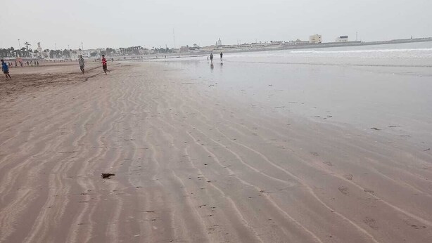 Ampla praia de areia com pegadas e algumas pessoas caminhando.
