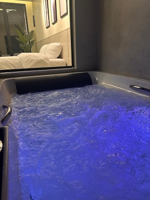 A hot tub with a blue light, a bed, and a mirror reflecting a plant and a wall with framed pictures. - Trio Chalet 1 (dead sea)