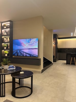 A modern living room with a large flat-screen TV, a small round table, and a staircase leading to a kitchen. - Trio Chalet 1 (dead sea)