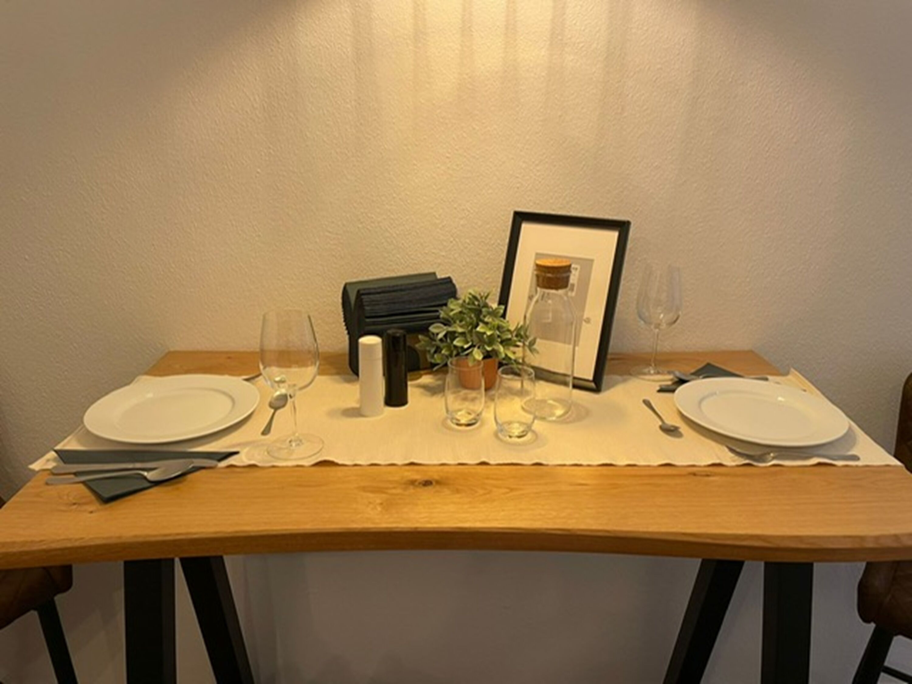 Ein Esstisch gedeckt mit Tellern, Besteck, Gläsern, einer kleinen Pflanze, einem gerahmten Bild und einem Kerzenständer.
