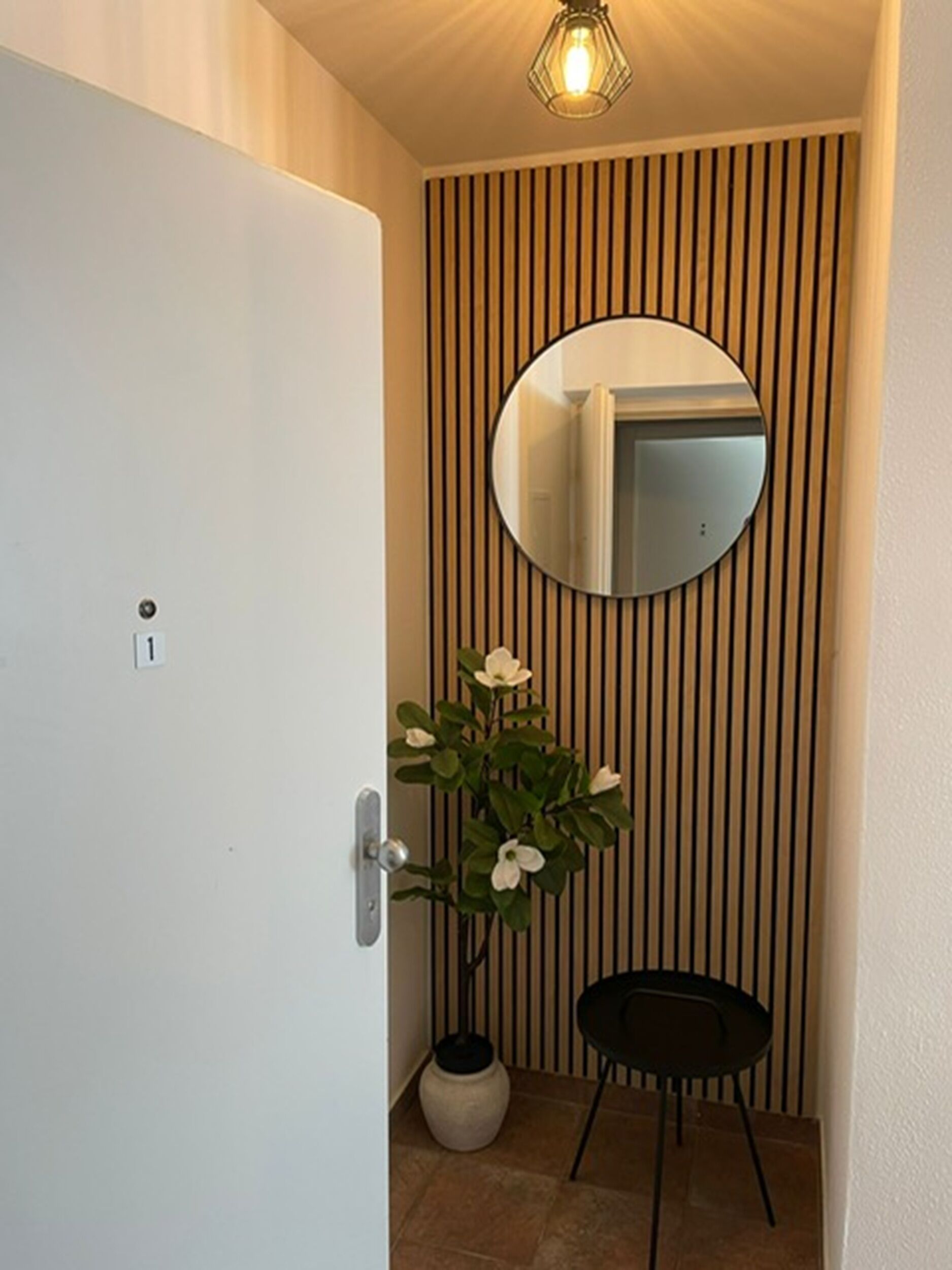 A door with a number plate, a round mirror, a potted plant with white flowers, and a small black chair.
