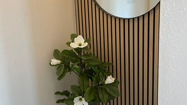 Ein wandmontierter Spiegel mit vertikalen Holzleisten, ein Blumentopf mit weißen Blüten und ein runder Spiegel.