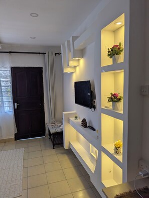 Ein modernes Wohnzimmer mit einer dunkelholzigen Tür, einem weißen Sofa, einem an der Wand befestigten Fernseher und einem eingebauten Regal mit Dekorationsobjekten.