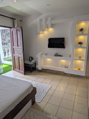 Ein Schlafzimmer mit einem Bett, einem an der Wand befestigten Fernseher, einem kleinen Tisch und einem Regal mit Blumen.
