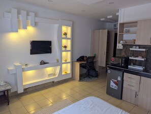 Ein modernes Zimmer mit Flachbildfernseher, eingebauten Regalen mit Blumen, Schreibtisch mit Stuhl, einem Bett, einer Küchenzeile und einem Kleiderschrank.
