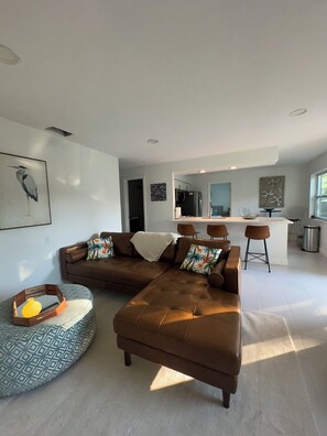 Uma sala de estar moderna com um sofá seccional, uma área de jantar com banquetas e uma cozinha ao fundo.