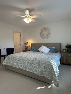 Quarto com cama, ventoinha no teto, um objeto decorativo na parede e mesa de cabeceira com abajur.