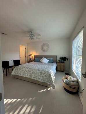 Ein Schlafzimmer mit einem Bett, einem Deckenventilator, einer Wanddekoration, einem Nachttisch mit Lampe und einem Fenster mit Jalousien.