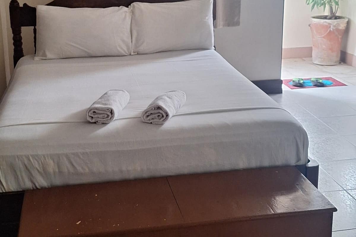 Una cama bien hecha con almohadas y toallas enrolladas, un cabecero de madera y una planta en maceta junto a la ventana.