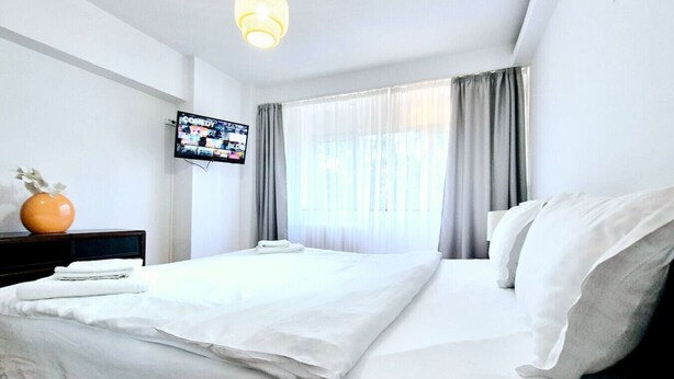 A hotel room with a bed, a TV on the wall, a window with curtains, and a hanging light fixture.
