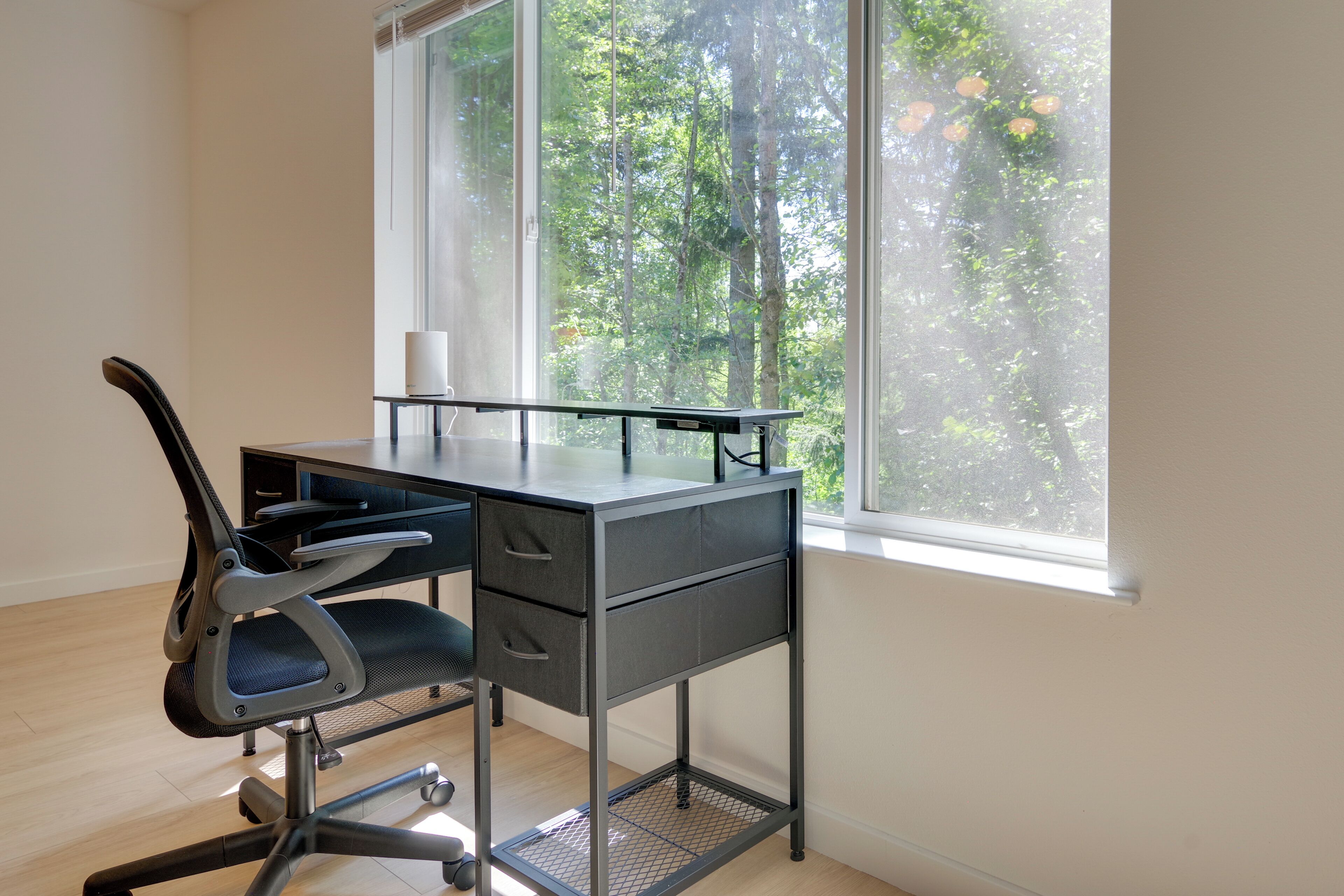A modern office desk with a chair, a window with a view of trees, and a white wall.